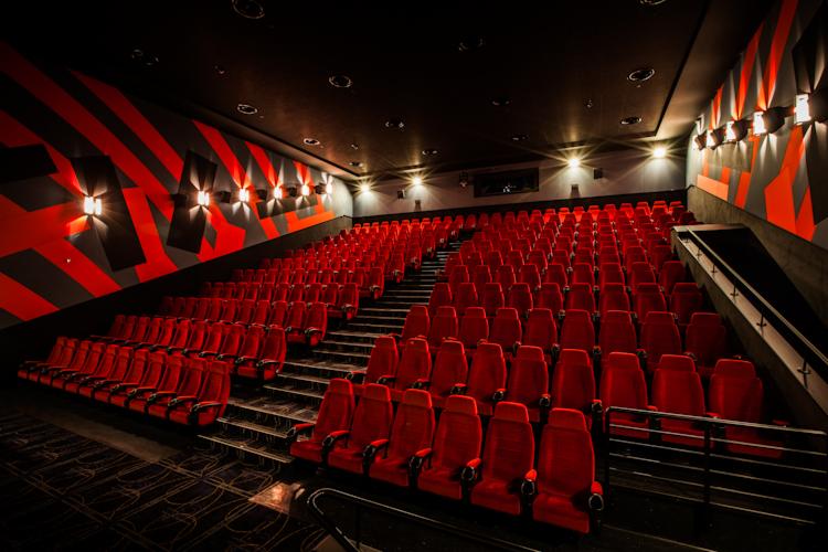 Wnętrze sali kinowej z rzędami czerwonych foteli, ciemną podłogą w geometryczne wzory i ścianami w odcieniach szarości z czerwonymi pasami oraz rzędem kinkietów.