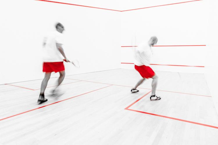 Dwóch mężczyzn w czerwonych spodenkach i białych koszulkach gra w squasha w białym pomieszczeniu z czerwonymi liniami, dynamiczne ujęcie z efektem rozmycia ruchu.