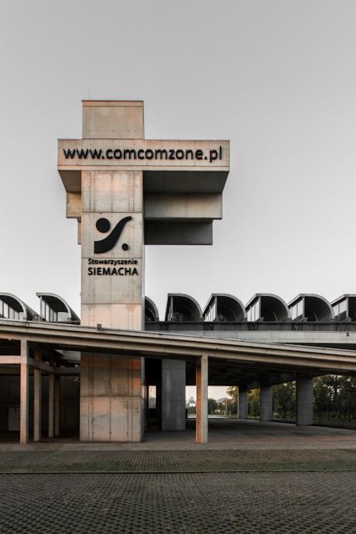 Betonowa konstrukcja budynku z napisem www.comcomzone.pl oraz logo Stowarzyszenia SIEMACHA na tle szarego nieba i częściowo zadaszonego placu.