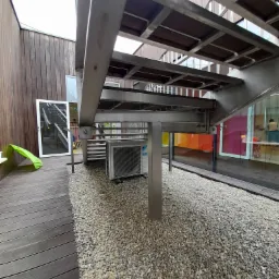 Zewnętrzna jednostka klimatyzacji Daikin pod metalowymi schodami na żwirowym podłożu, widoczna z poziomu gruntu, z fragmentem drewnianego tarasu i kolorowymi oknami w tle.