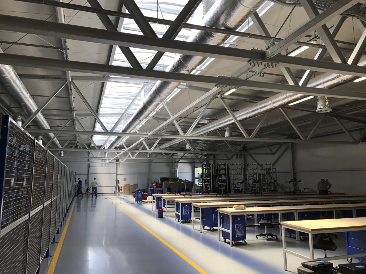 Wnętrze hali produkcyjnej z widocznymi kanałami wentylacyjnymi pod sufitem, rzędami stołów warsztatowych i pracownikami w tle.