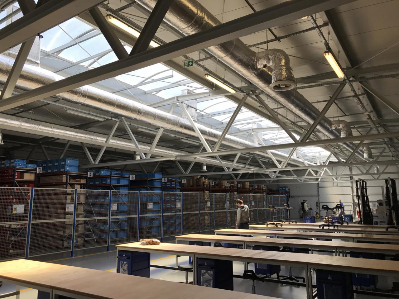 Wnętrze hali produkcyjnej z widoczną instalacją wentylacyjną pod sufitem, oświetleniem i regałami magazynowymi w tle.