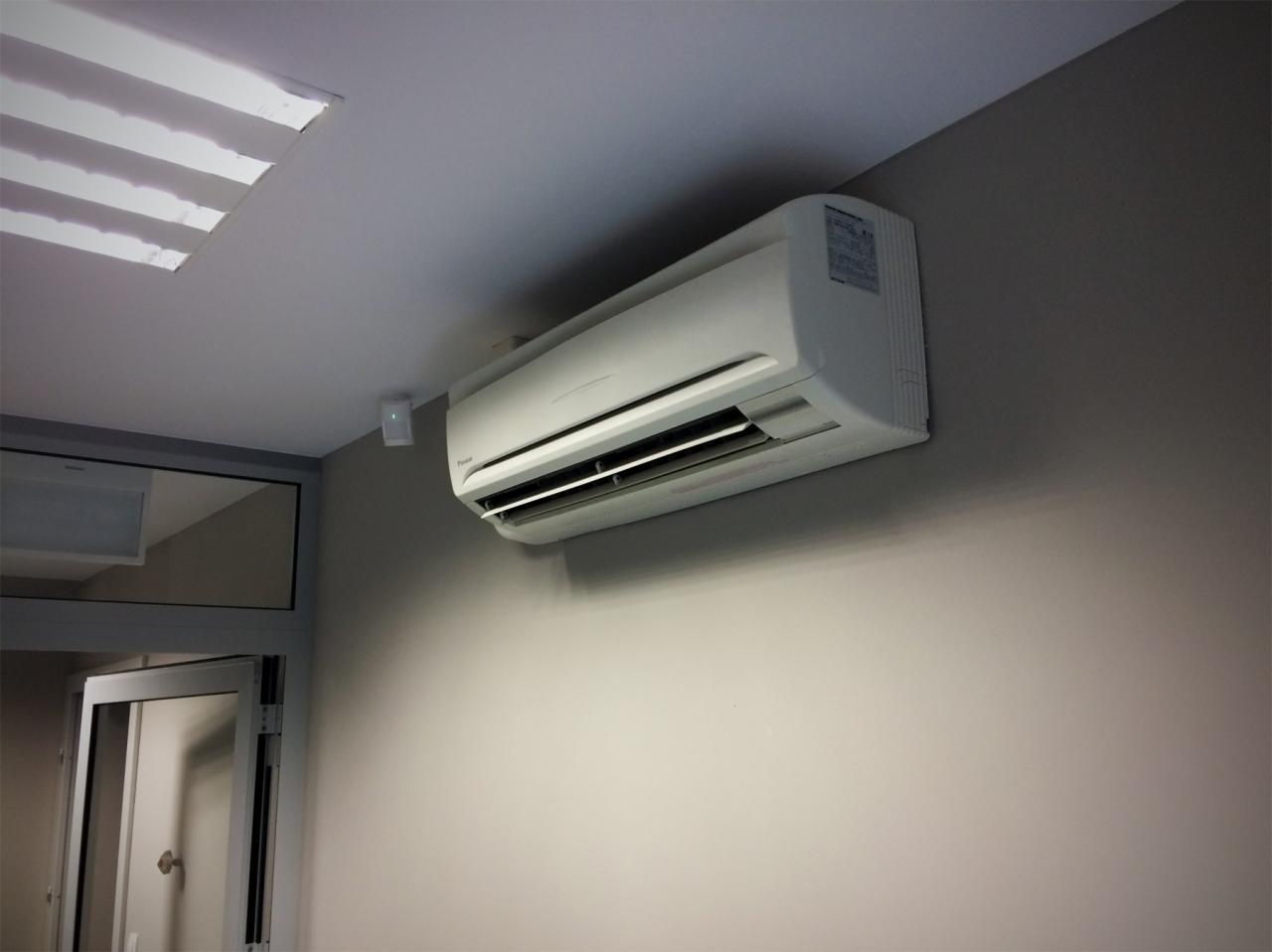 Biała klimatyzacja typu split zamontowana na szarej ścianie w pomieszczeniu biurowym, widoczna lampa sufitowa i fragment przeszklonych drzwi wejściowych.