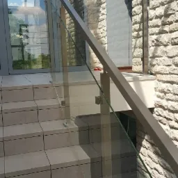 Szklana balustrada z metalowymi słupkami i poręczą zamontowana przy schodach zewnętrznych, z widoczną kamienną elewacją budynku w tle i dekoracyjnymi balonami przy wejściu.