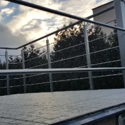 Balkon z nowoczesną, metalową konstrukcją zabezpieczającą, widok na pochmurne niebo i zielone drzewa w tle.