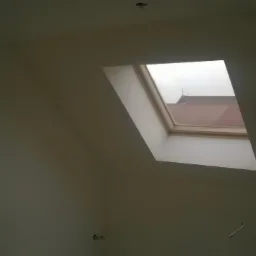 Ściana w odcieniach szarości z oknem dachowym; widok na dach sąsiedniego budynku. Świeżo pomalowane wnętrze poddasza, jasne światło dzienne.