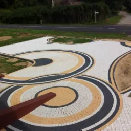 Ogród z dekoracyjną nawierzchnią z kostki brukowej w odcieniach bieli, beżu i czerni, ułożonej w koncentryczne okręgi i łuki, z elementami drewnianej architektury ogrodowej w tle oraz fragmentem...