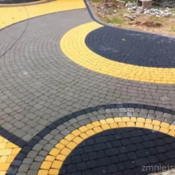Ścieżka ułożona z kostki brukowej w odcieniach szarości, czerni i żółci, tworząca dekoracyjny wzór łuków i okręgów, z widocznym fragmentem terenu w tle.