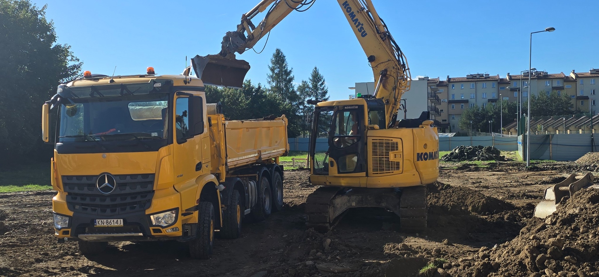Żółta koparka Komatsu ładuje ziemię do wywrotki Mercedes na placu budowy. W tle budynki mieszkalne i błękitne niebo.