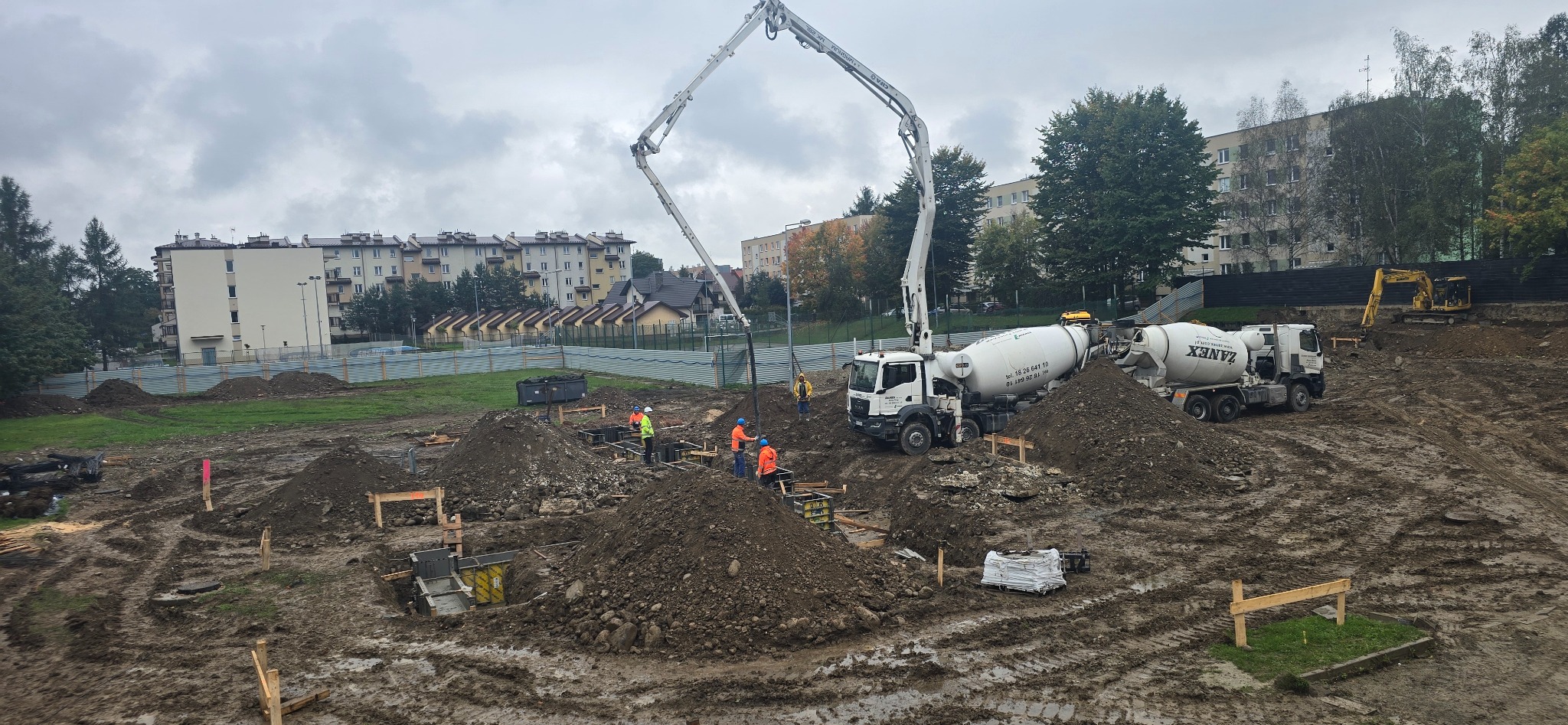 Wylewanie betonu do szalunków fundamentowych na placu budowy. Widoczne betoniarki, pompa do betonu i pracownicy w odblaskowych ubraniach. W tle budynki mieszkalne.