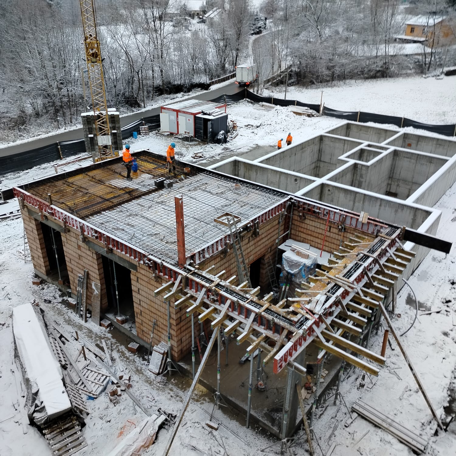 Zimowy kadr z budowy domu: fundamenty, ściany z cegły, zbrojenie stropu. Dwóch robotników w pomarańczowych ubraniach na stropie. Żuraw budowlany w tle.