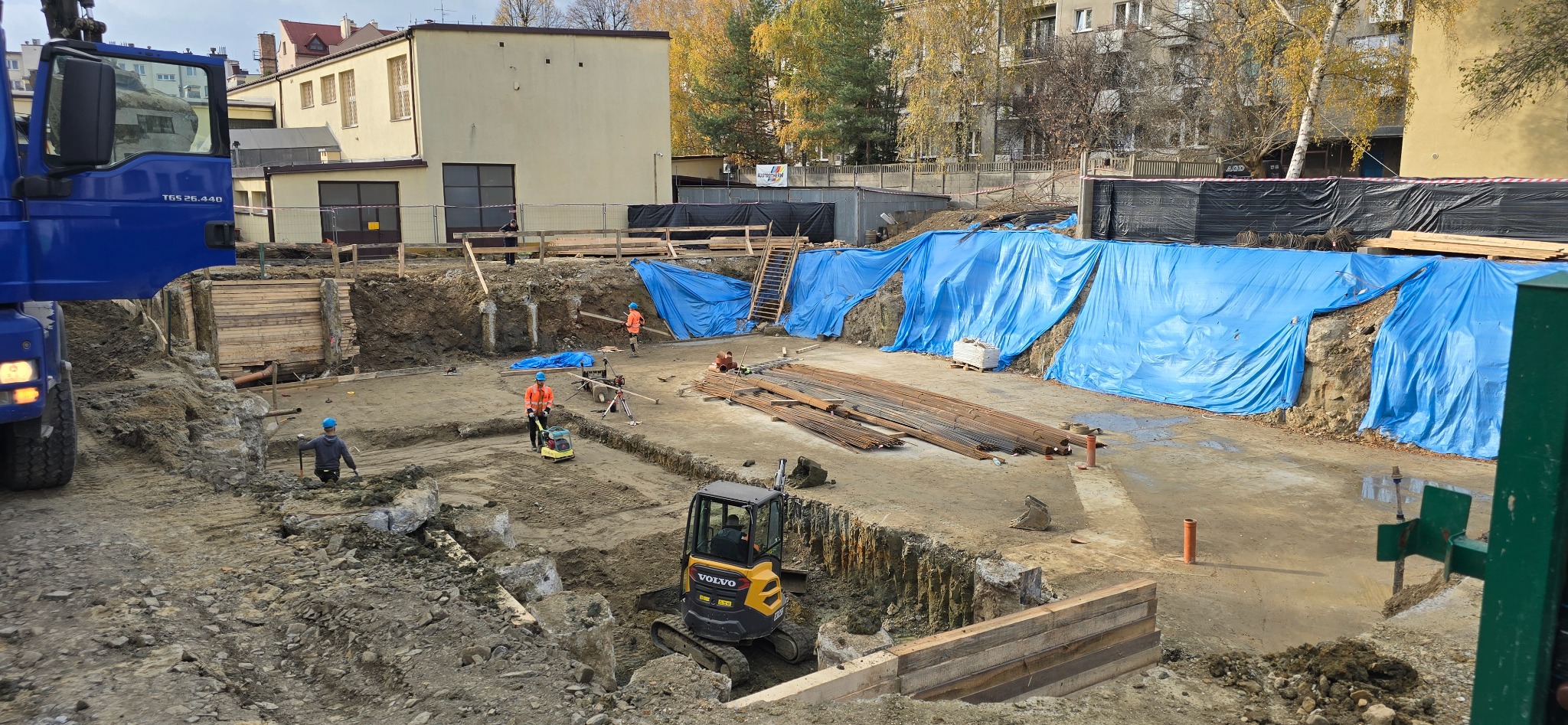 Wykonanie wykopu pod fundamenty z użyciem koparki Volvo. Widoczne zabezpieczenia wykopu, pracownicy w odblaskowych ubraniach i przygotowane zbrojenie.