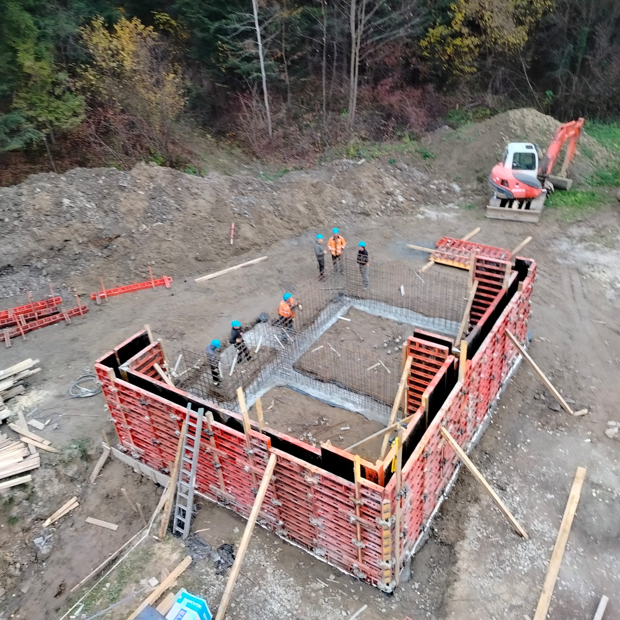 Z góry widoczny wykop fundamentu z deskowaniem i zbrojeniem. Na budowie ekipa w kaskach i mała koparka. Jesienne drzewa w tle.