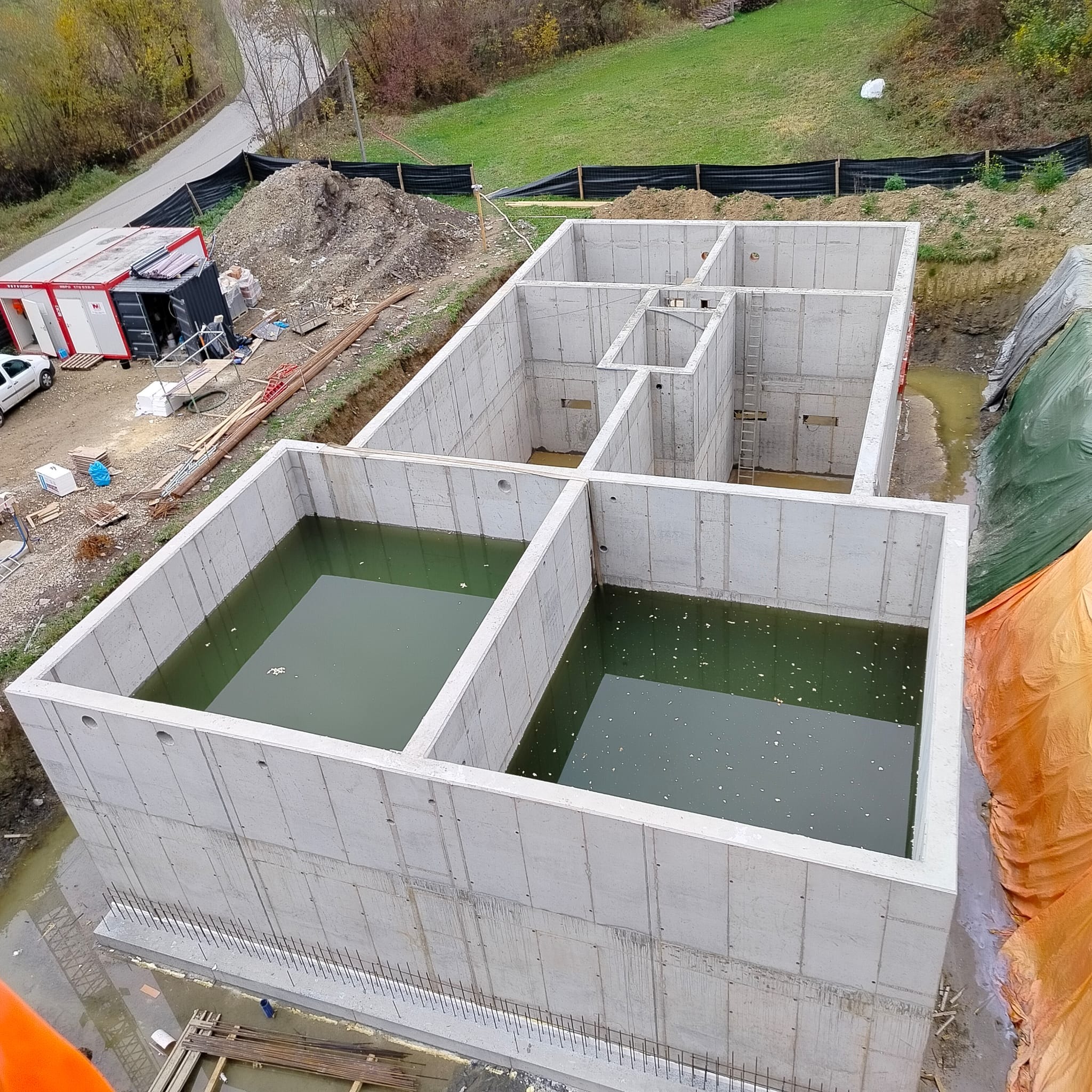 Żelbetowy fundament z wodą wewnątrz, widok z góry. Widoczne ślady po szalunkach, wystające pręty zbrojeniowe i drabina wewnątrz konstrukcji.