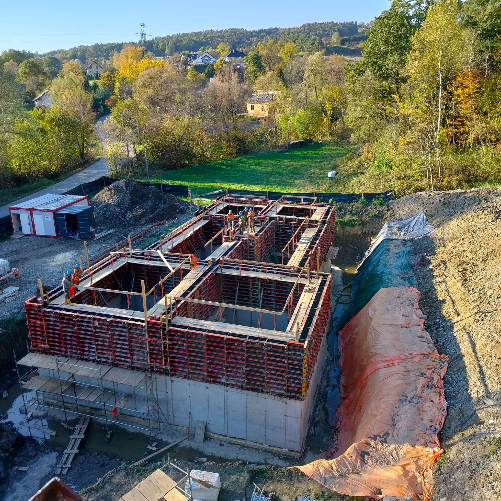 Wysoka perspektywa na plac budowy z szalunkami fundamentów, pracownikami w kaskach i otaczającym krajobrazem z drzewami w jesiennych barwach.