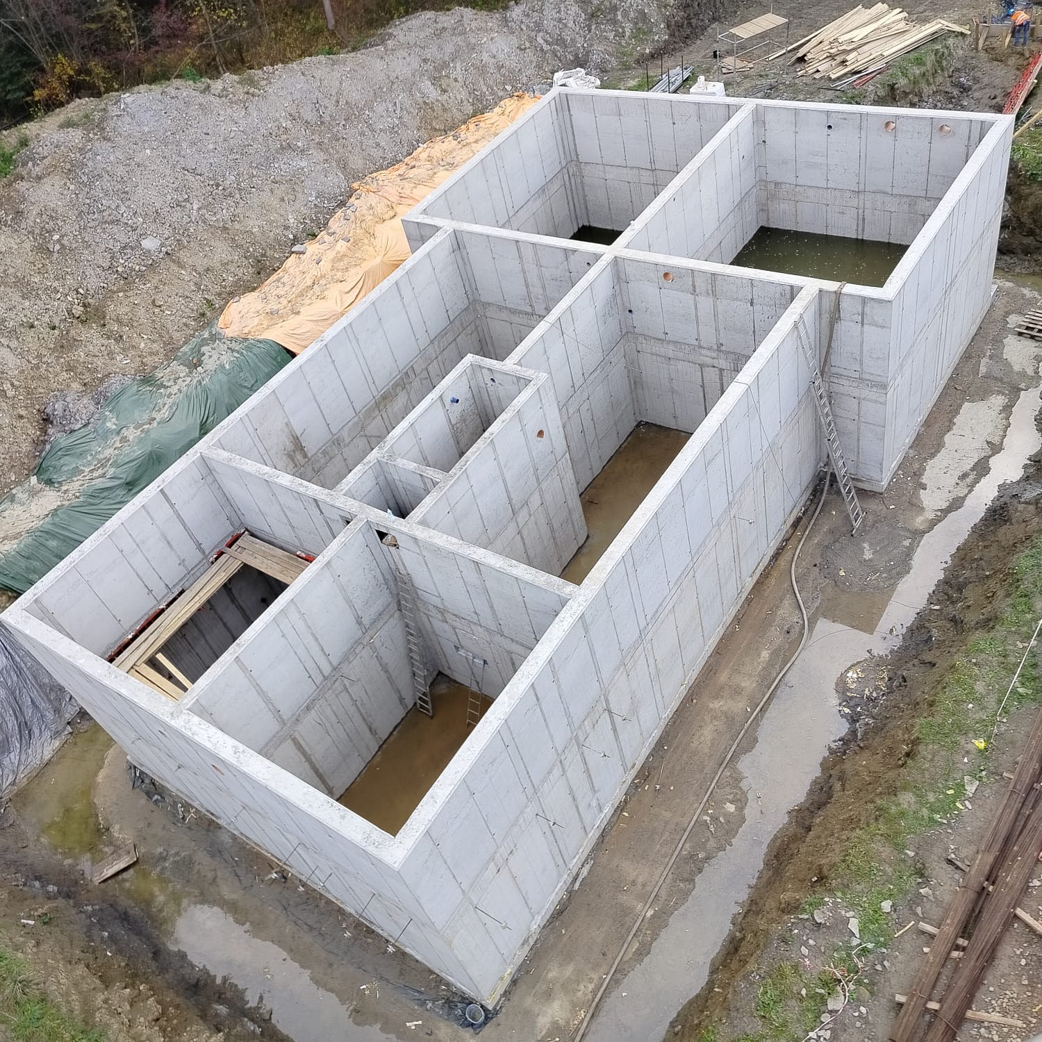 Surowy stan fundamentów z betonu komórkowego, widok z góry. Wnętrze zalane wodą, drabina oparta o ścianę. Teren budowy w jesiennej scenerii.