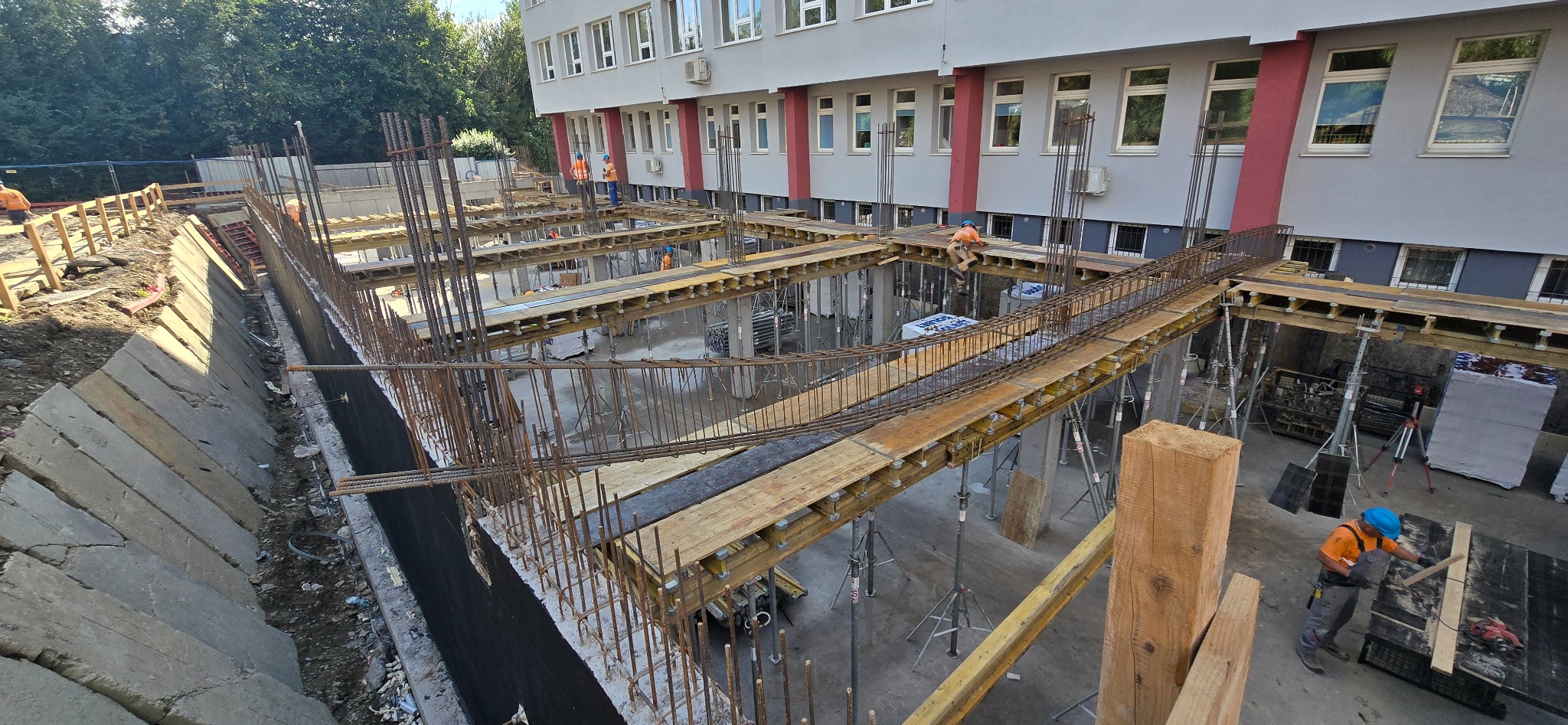 Wykonywanie szalunków i zbrojenia fundamentów budynku. Widoczne drewniane konstrukcje wsporcze, stalowe pręty zbrojeniowe i pracownicy w kaskach. Budynek w tle.