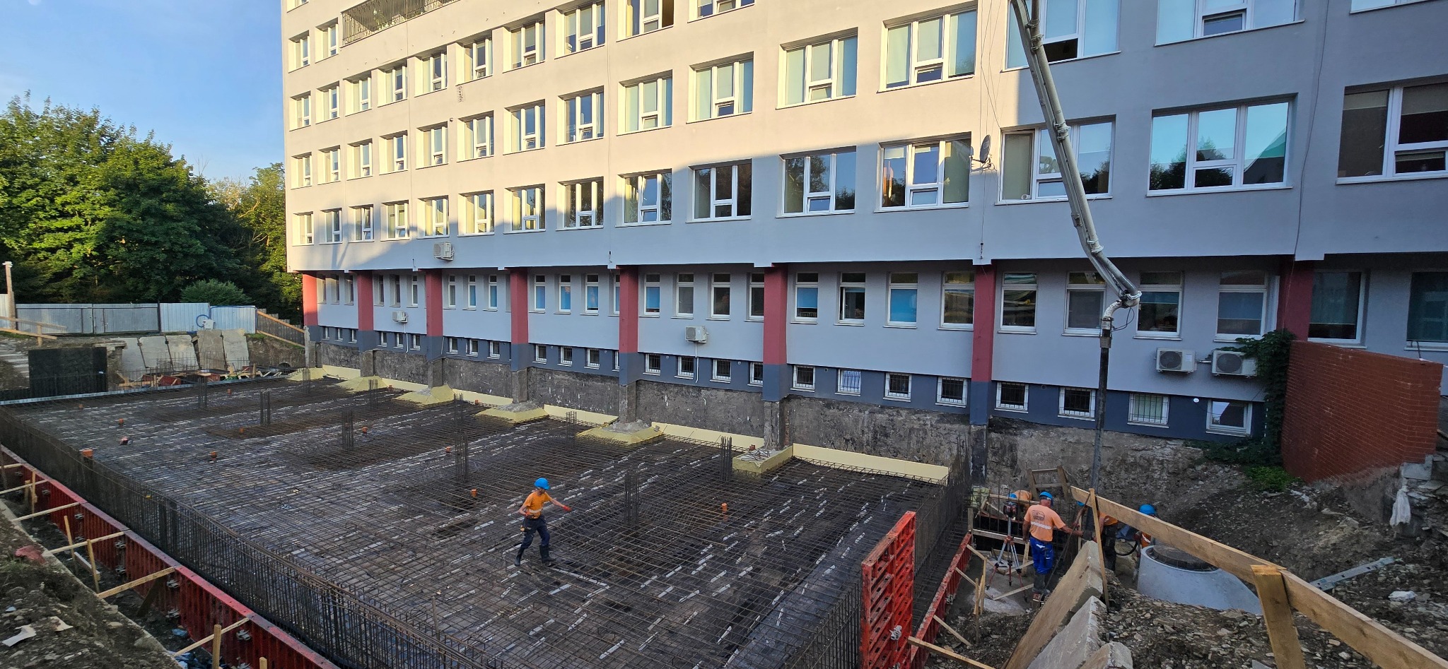 Zbrojenie fundamentów budynku z widoczną konstrukcją z prętów stalowych, pracownikami w kaskach i pompą do betonu. W tle budynek z licznymi oknami.