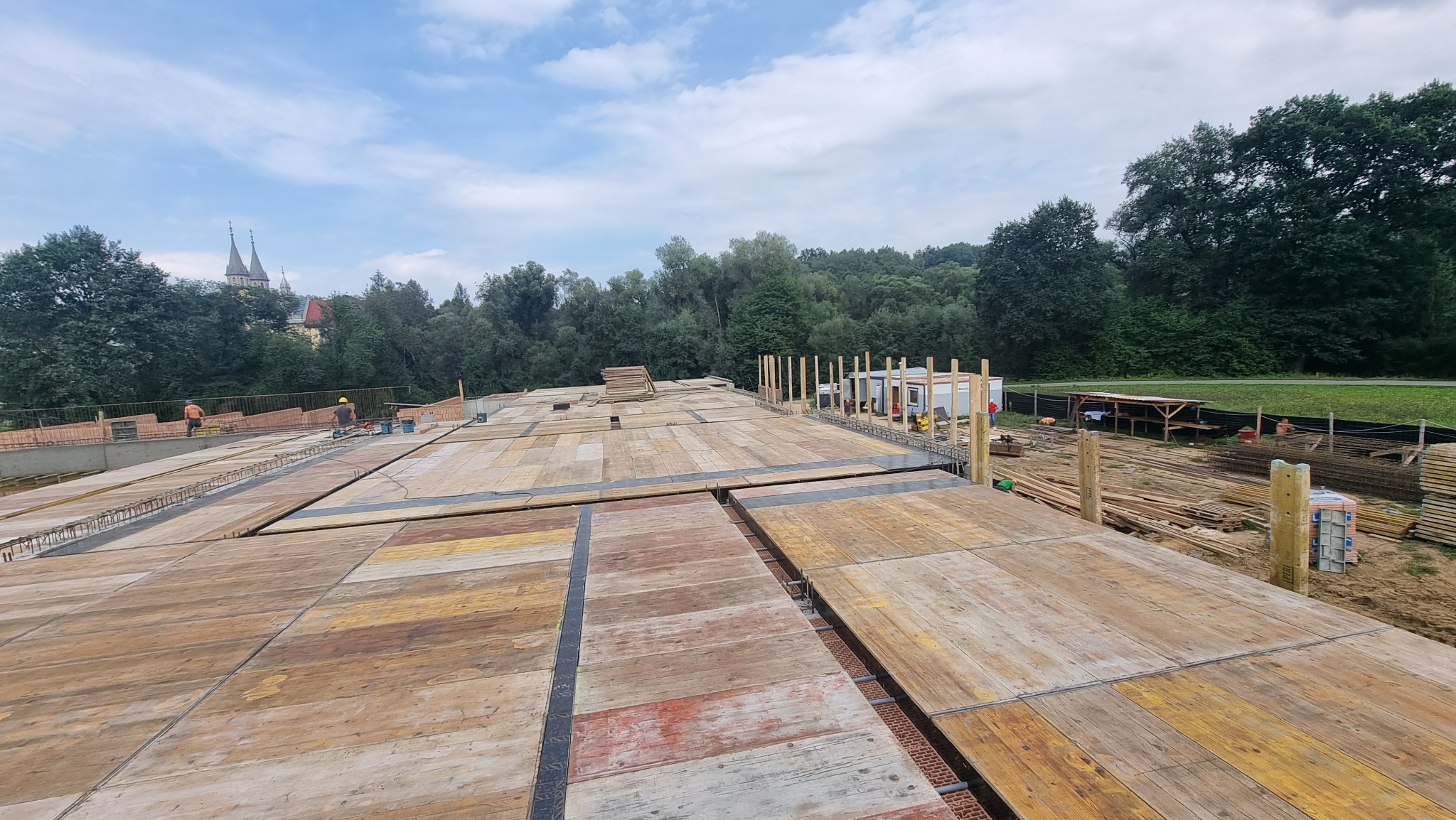 Zalana szalunkami płyta fundamentowa z widocznym zbrojeniem, przygotowana do wylania betonu. W tle budynek z wieżami i zieleń. Dwóch pracowników budowlanych w oddali.