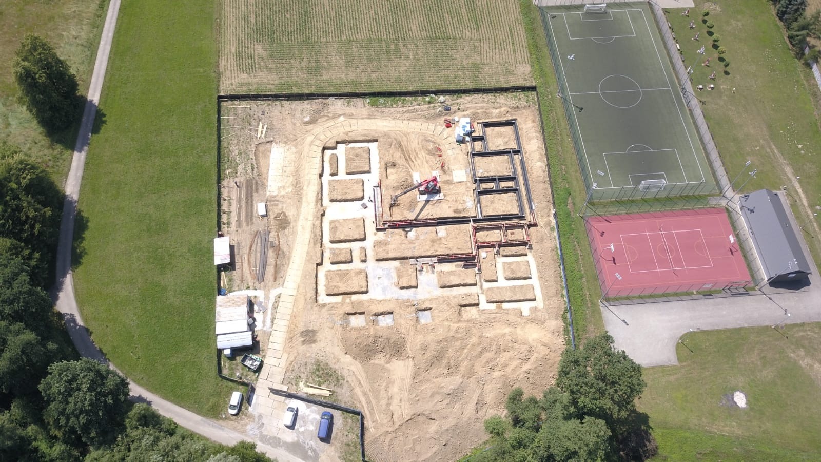 Widok z lotu ptaka na plac budowy z wykopami pod fundamenty, częściowo zbrojonymi, z dźwigiem w centrum. W tle boiska sportowe i tereny zielone.