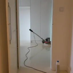 Błyszcząca, jasna podłoga po polerowaniu, widoczna maszyna polerska i butelka środka czyszczącego przy ścianie, ujęcie zza otwartych drzwi.