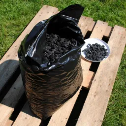 Czarny worek wypełniony brykietami na drewnianej palecie, obok biały talerz z brykietami, na tle zielonej trawy.