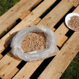 Pellet drzewny w przezroczystym worku i na białym talerzu, ułożony na drewnianej palecie na trawie.