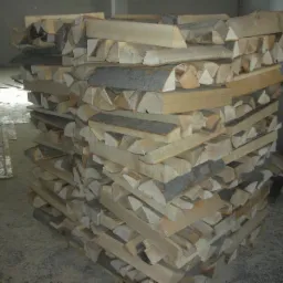 Ułożony stos drewna bukowego, kawałki o różnej wielkości, widoczne sęki i kora, szare tło betonowej podłogi i ściany.