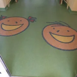 Zielona podłoga z wykładziny PCV w przedszkolu, z wtopionymi w strukturę podłogi wizerunkami uśmiechniętych dziecięcych twarzy, chłopięcej i dziewczęcej.