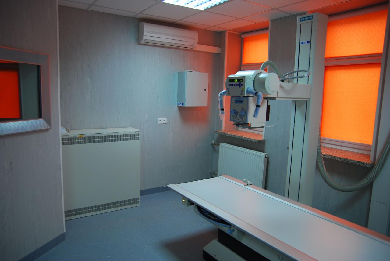 Szare pomieszczenie z niebieską podłogą, wyposażone w biały aparat rentgenowski firmy Siemens, z pomarańczowymi roletami w oknach i klimatyzacją na ścianie.
