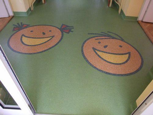 Zielona podłoga z wykładziny PCV w przedszkolu, z wtopionymi w strukturę podłogi wizerunkami uśmiechniętych dziecięcych twarzy, chłopięcej i dziewczęcej.