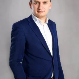 Portret biznesowy mężczyzny w niebieskiej marynarce i białej koszuli, na szarym tle. Widoczne logo fotografa w dolnej części zdjęcia.