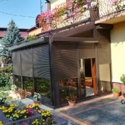 Zadaszony taras z brązowymi roletami zewnętrznymi, otoczony kolorowym ogrodem i kwiatami w donicach na balkonie domu.