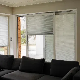 Jasne, plisowane osłony okienne w nowoczesnym salonie z szarą sofą i widokiem na patio.