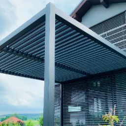 Nowoczesna pergola z regulowanymi lamelami na drewnianym tarasie, z widokiem na zielony krajobraz w tle i fragmentem domu z żaluzjami zewnętrznymi.