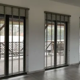 Wnętrze z trzema oknami przesłoniętymi drewnianymi żaluzjami 50 mm w odcieniach szarości, widok na drewniany taras z balustradą.