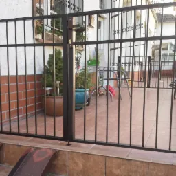 Czarne metalowe ogrodzenie z pionowymi prętami, widok na taras z meblami ogrodowymi i rowerem. Ceglana ściana budynku w tle.