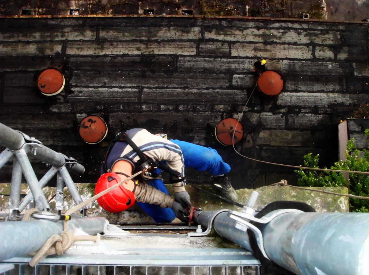 Pracownik w kasku, na wysokości, zabezpieczony linami, wykonuje prace na elewacji budynku z cegły z widocznymi, okrągłymi elementami wentylacyjnymi.