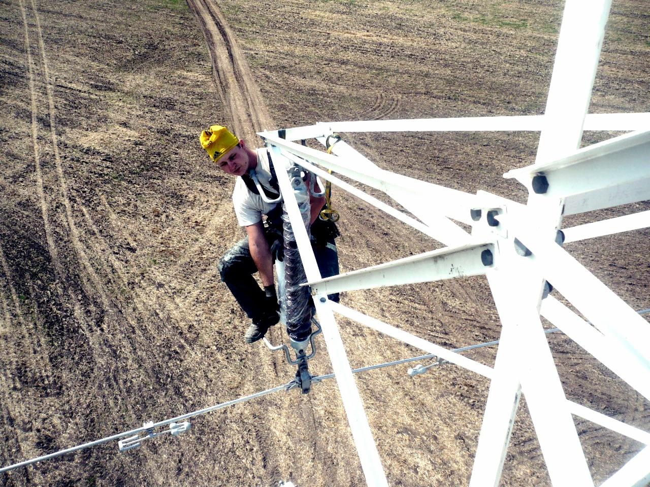 Pracownik w żółtym kasku, zabezpieczony linami, wspina się po konstrukcji wieży energetycznej. Widok z góry na pole i drogę polną.