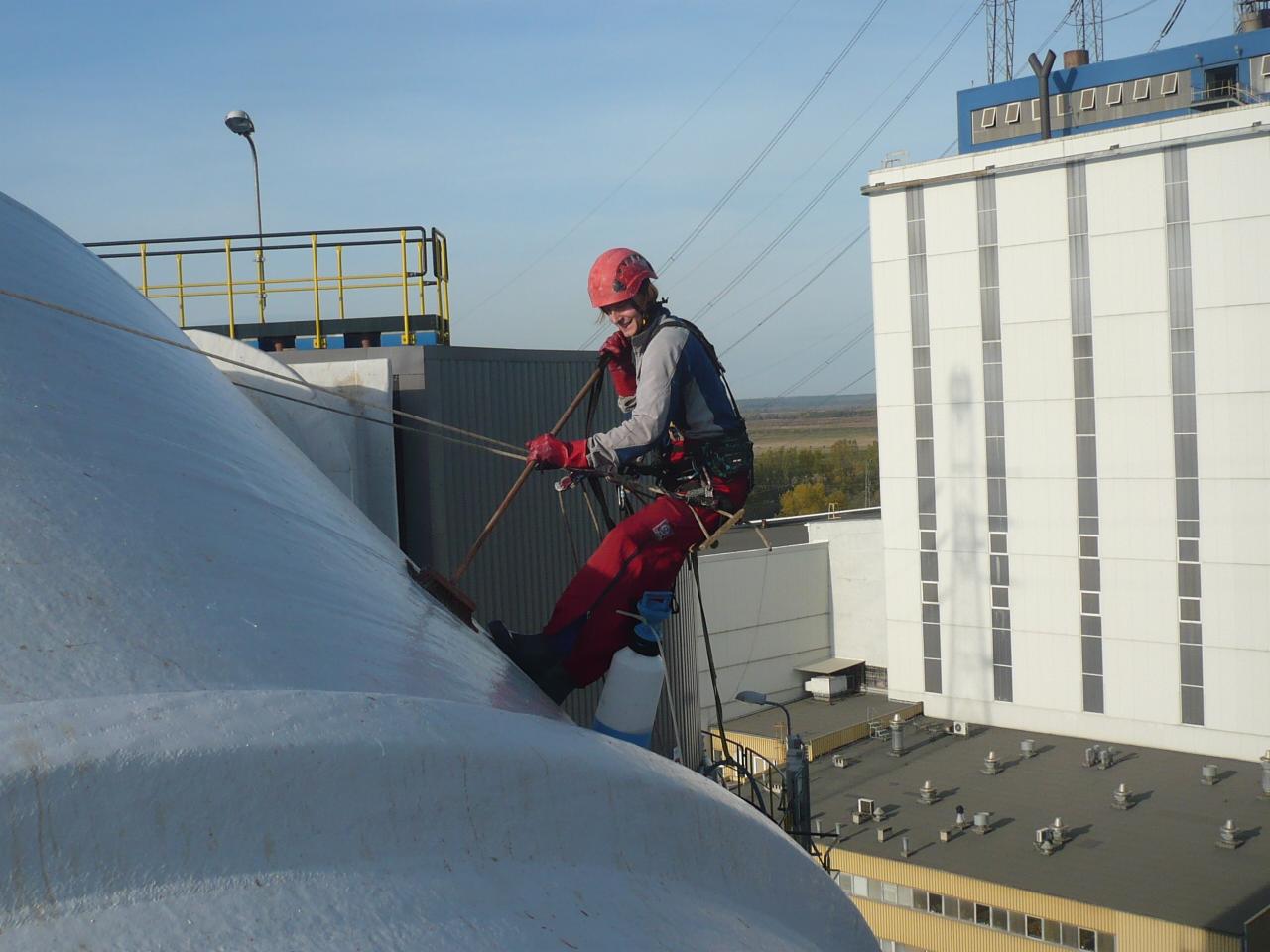 Pracownik w kasku i kombinezonie czyści kopułę budynku przemysłowego, zabezpieczony linami. Widok z góry na dachy budynków i elementy infrastruktury.