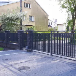 Automatyczna brama przesuwna z metalowymi sztachetami, zamontowana na słupkach z szarego kamienia łupanego, widok z zewnątrz posesji na tle domu z kwitnącym drzewem.