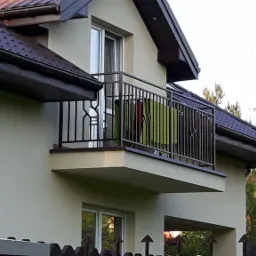 Metalowa balustrada balkonowa w kolorze ciemnego brązu, z widocznymi pionowymi prętami i poziomymi poręczami, na tle elewacji domu z brązowym dachem i fragmentem ogrodzenia.