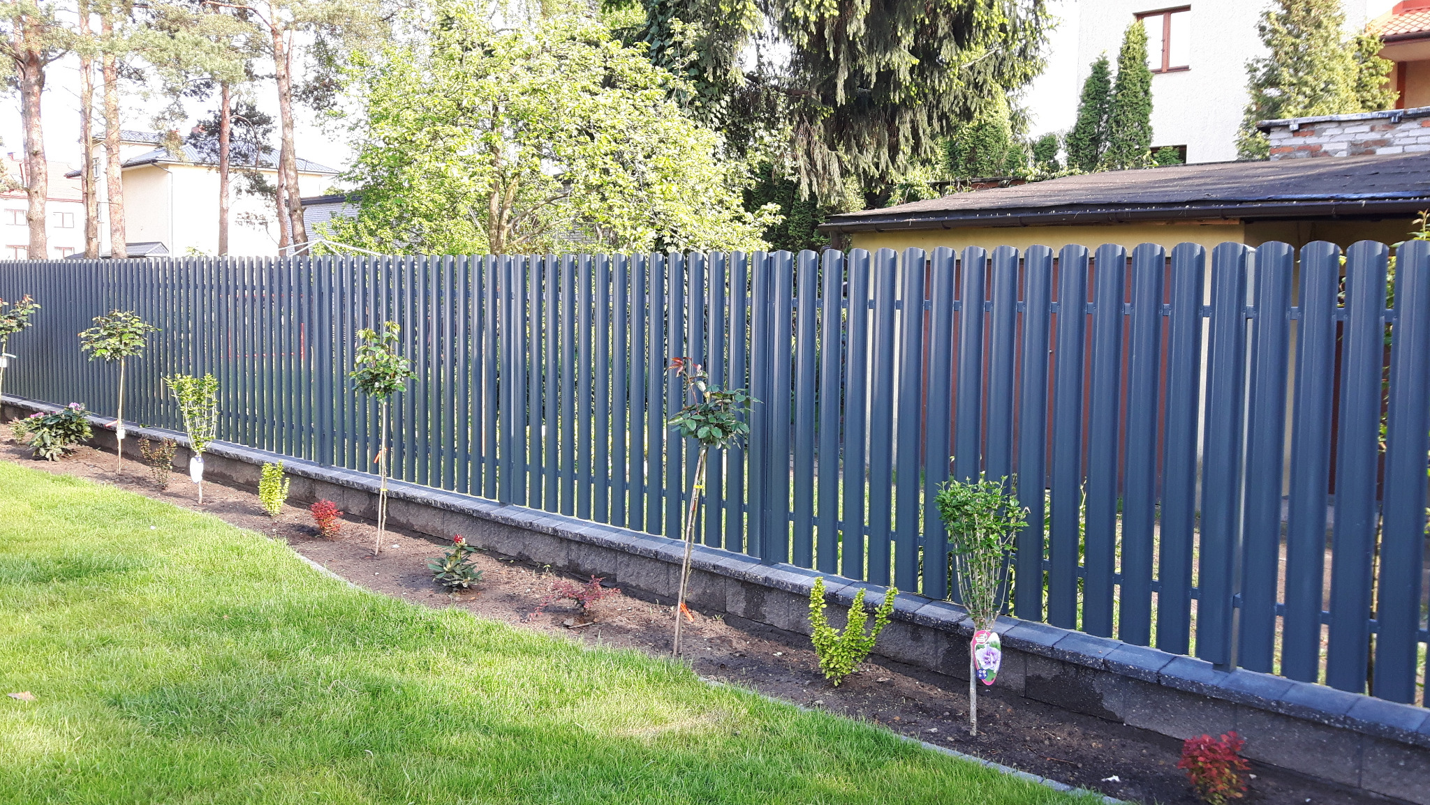 Nowoczesne, szare ogrodzenie z pionowych paneli, z niską podmurówką i świeżo posadzoną roślinnością, na tle zielonego trawnika i zabudowań mieszkalnych.