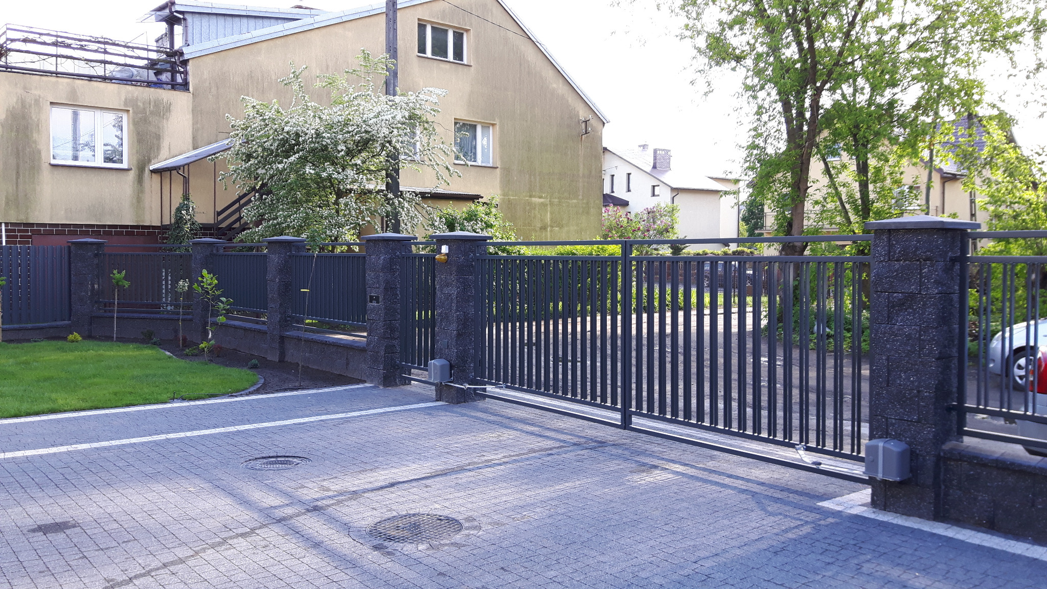 Automatyczna brama przesuwna z metalowymi sztachetami, zamontowana na słupkach z szarego kamienia łupanego, widok z zewnątrz posesji na tle domu z kwitnącym drzewem.