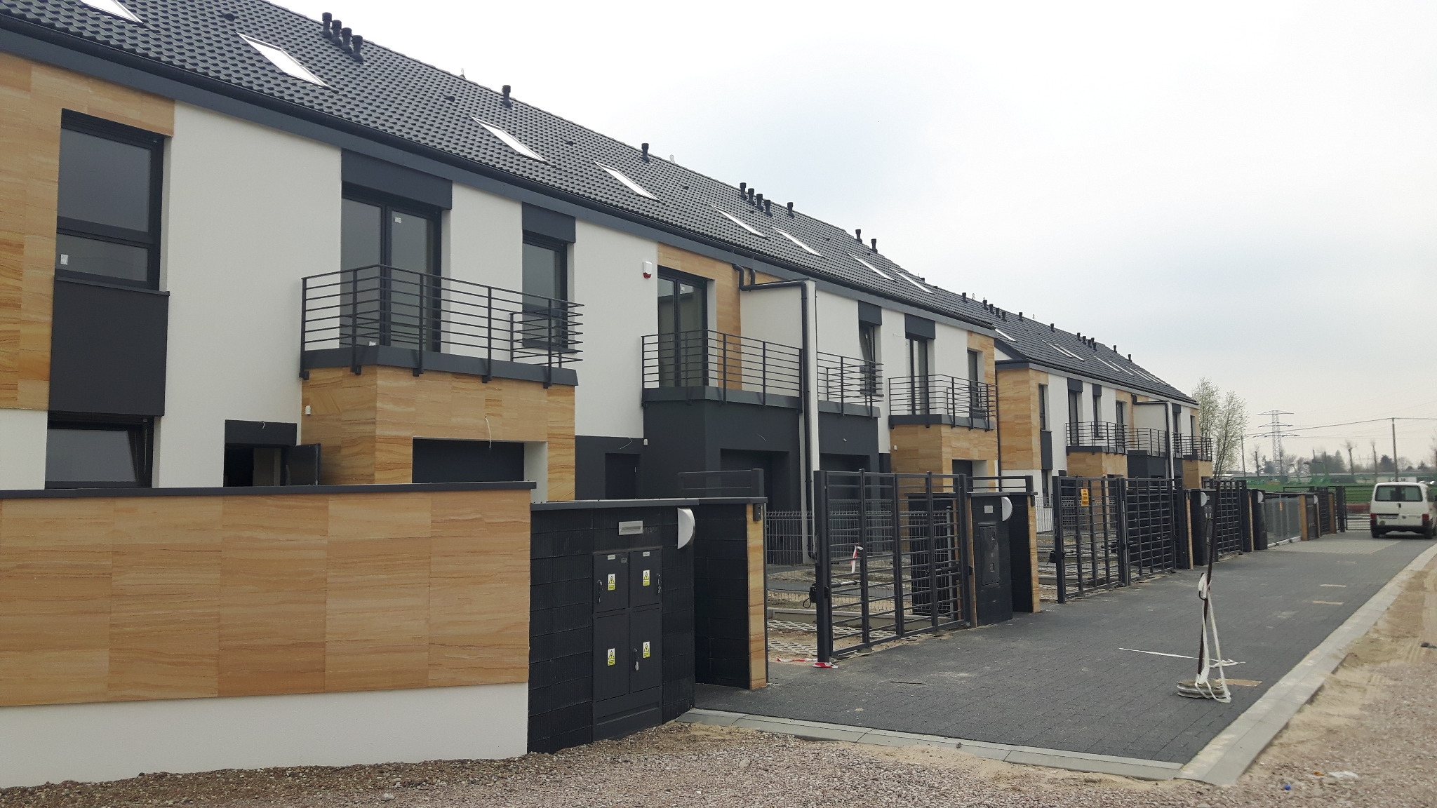 Szereg nowo wybudowanych domów szeregowych z czarnymi, metalowymi balustradami balkonowymi i czarnymi bramami wjazdowymi, widok z poziomu ulicy.