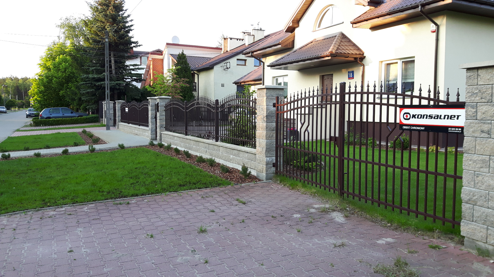 Ozdobna brama i przęsła z metalu w kolorze ciemnego brązu, zamontowane na podmurówce z szarego kamienia łupanego, z tabliczką informacyjną 'Obiekt Chroniony'.