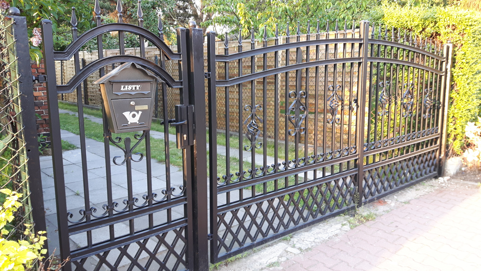 Otwarta, czarna, metalowa brama wjazdowa z dekoracyjnymi elementami i skrzynką na listy, widoczna na tle ceglanego muru i zieleni.