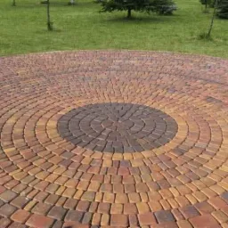 Okrągła nawierzchnia z kostki brukowej w odcieniach brązu i pomarańczy, ułożona w koncentryczne kręgi, otoczona zielonym trawnikiem z drzewami w tle.