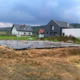 Zalane fundamenty domu jednorodzinnego na działce, widoczne zbrojenia, folia i wystające słupki. W tle zabudowa jednorodzinna i pochmurne niebo.