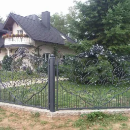 Kuta, metalowa, dekoracyjna brama w kolorze grafitowym, z motywem roślinnym, na tle domu z balkonem i ogrodem. Podmurówka z kamienia.
